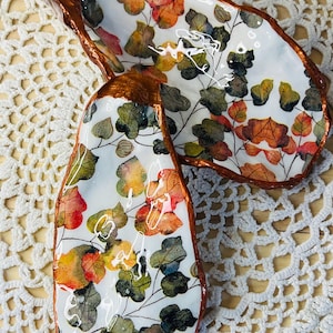May include: Two decorative oyster shell dishes. The interior has a white background with colourful leaf patterns in red, orange, yellow, and green. The edges are trimmed with a metallic copper-coloured material. The dishes are on a white lace surface.