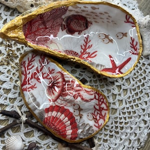 Könnte beinhalten: Zwei dekorative Austernmuscheln mit goldfarbenem Rand. Die Muscheln sind mit einem weißen Hintergrund und roter Koralle, Fischen und Muscheln bemalt.