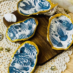 May include: Four oyster shell dishes with gold rims and marine-themed designs. Each shell features a unique blue and white illustration of sea creatures and plants. The shells are arranged on a wooden board and a white lace doily.