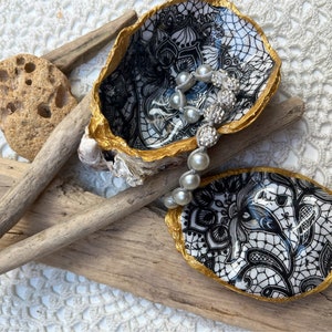 Pode incluir: Dois pratos de concha de ostra com borda dourada e um padrão de renda branco e preto. Um colar de pérolas está descansando em um dos pratos.