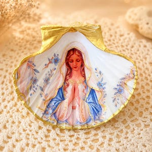 Puede incluir: Una concha marina decorativa con una ilustraci&oacute;n pintada a mano de una figura de cabello largo, con un velo blanco y una t&uacute;nica azul, con las manos juntas en oraci&oacute;n. La concha tiene un borde pintado en oro y detalles florales azules.