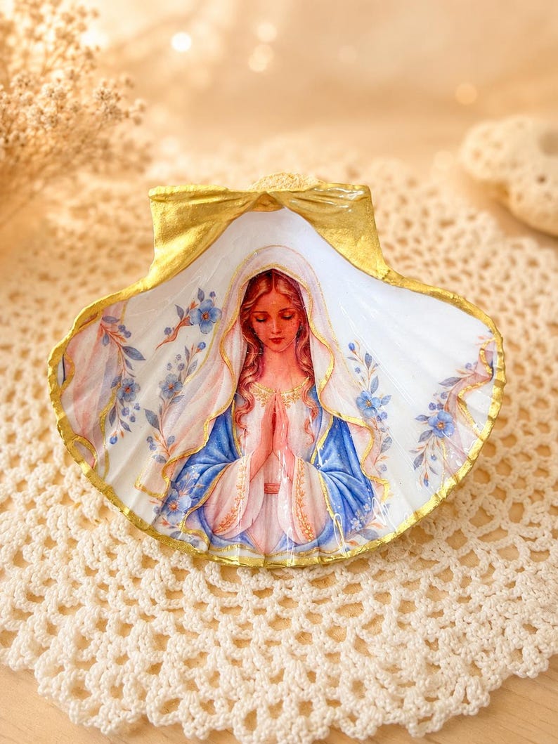 Puede incluir: Una concha marina decorativa con una ilustraci&oacute;n pintada a mano de una figura de cabello largo, con un velo blanco y una t&uacute;nica azul, con las manos juntas en oraci&oacute;n. La concha tiene un borde pintado en oro y detalles florales azules.