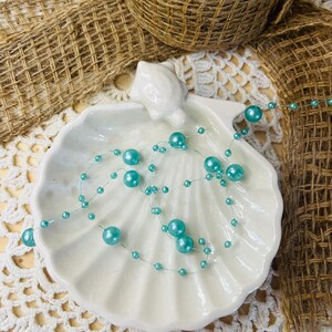 May include: A white ceramic seashell-shaped dish with a string of turquoise beads. The beads are of varying sizes and are strung together on a thin, clear wire. The dish is on a white lace doily, with burlap ribbon and a woven basket in the background.