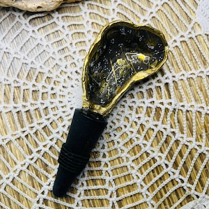 May include: A gold and black bottle stopper shaped like a seashell with a gold-painted interior and a black rubber stopper. The seashell has a design of gold stars and swirls on a black background.