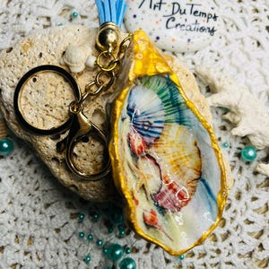 May include: A gold-painted oyster shell keychain with a colorful painted design inside. The keychain has a blue tassel and a gold chain with a ring.