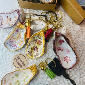 May include: A set of six oyster shell keyrings with gold trim and colourful painted designs. Each keyring features a different design and includes a gold key ring and a tassel. The text "Fabrication Artisanale" is visible on a tag.