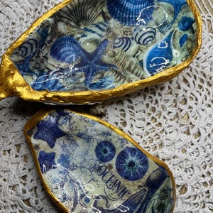 Könnte beinhalten: Zwei dekorative Keramikschalen mit nautischem Thema. Die größere Schale ist goldfarben mit einem blauen und weißen Muschelmotiv. Die kleinere Schale ist goldfarben mit einem blauen und weißen Krakenmotiv und dem Text "Oceanic" und "Better in the Water".