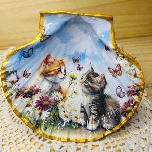 Könnte beinhalten: Eine Muschelschalen-Schale mit goldener Einfassung, die eine Aquarellmalerei von zwei Kätzchen auf einer Wiese mit Gänseblümchen und Schmetterlingen zeigt. Die Kätzchen schauen sich an.