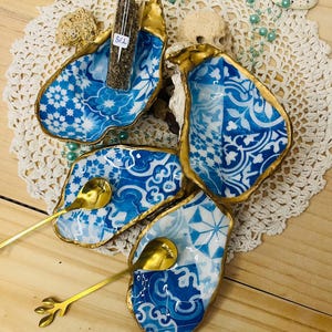 May include: A set of four blue and white patterned oyster shell dishes with gold rims, each holding a small gold spoon. A small glass vial labeled "SEL" sits in one shell. The shells rest on a white lace doily.