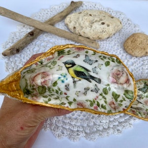 Peut inclure: Une coquille de coquillage blanche avec un bord doré, décorée d'un motif floral et d'un petit oiseau. La coquille est un objet décoratif.