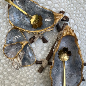 Puede incluir: Dos platos de concha de ostra grises y dorados con cucharas doradas en forma de flor. Los platos est&aacute;n sobre una superficie de crochet blanco.