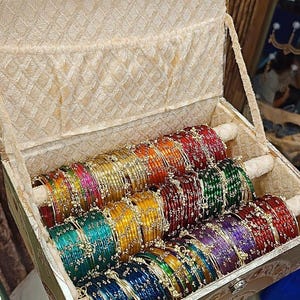 May include: A decorative box filled with colorful bangles. The bangles are arranged in rows and come in various colors, including red, green, yellow, and blue. The box has a patterned exterior and a soft, cream-colored interior.