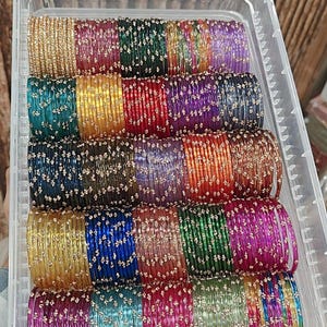 May include: A clear plastic container filled with stacks of colorful bangles. The bangles are in various colors, including gold, green, red, purple, and blue, and are adorned with small, sparkling embellishments. The bangles are a traditional jewelry item.