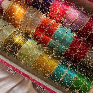 May include: A selection of vibrant glass bangles in a decorative box. The bangles are in shades of orange, yellow, green, teal, red, pink, and purple. Each bangle is decorated with small gold-coloured beads. The box has a pink and gold trim.