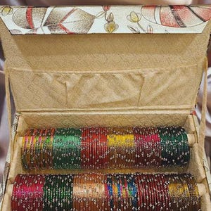 May include: A box of colourful bangles, with rows of red, green, yellow, and orange bangles. The box is beige with a floral pattern on the lid. The bangles are displayed inside the open box.