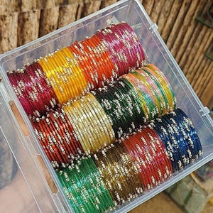 Puede incluir: Un recipiente de plástico transparente lleno de pilas de pulseras coloridas. Las pulseras son en tonos de rojo, naranja, amarillo, verde y azul, con adornos plateados. Las pulseras están apiladas ordenadamente dentro del recipiente.