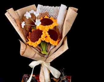 Puede incluir: Un ramo de girasoles de ganchillo con centros marrones y pétalos amarillos, envuelto en papel de periódico. El ramo está atado con una cinta blanca y descansa sobre una pila de libros. También se incluyen flores blancas de acento.