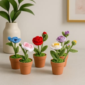 以下が含まれることがあります： 小さなテラコッタの鉢に入った、手作りのかぎ針編みの花のコレクション。花は青、ピンク、赤、白、紫など様々な色があり、緑色の茎と葉が付いています。鉢には緑色の苔が詰められています。