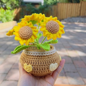 Pode incluir: Um arranjo de girassóis de crochê em um pequeno vaso marrom. Os girassóis têm pétalas amarelas brilhantes e centros marrons. O vaso é decorado com aplicações em forma de coração amarelo e uma borda de renda branca.
