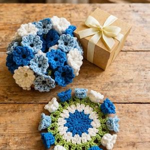 May include: A round crochet wreath featuring clusters of blue and white flowers, a circular crochet doily with a green, white, and blue floral pattern, and a square cardboard gift box tied with a cream-colored ribbon on a wooden surface.