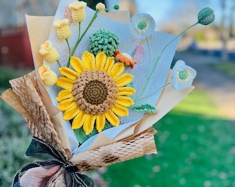 Puede incluir: Un ramo de flores de ganchillo, que incluye un girasol amarillo, flores de amapola blancas y flores amarillas en forma de campana. El ramo está envuelto en papel marrón y atado con una cinta negra.
