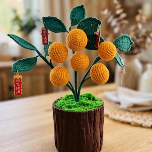 May include: A crocheted orange tree with five round orange fruits and dark green leaves. The tree is planted in a brown, textured pot with green moss-like material on top. Two small red rectangular charms with gold text hang from the branches.