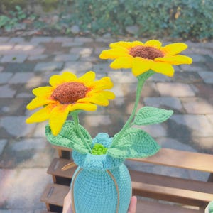 May include: Crocheted sunflowers with yellow petals and brown centers in a turquoise vase. The vase has a drawstring closure and is filled with green leaves. The sunflowers are handmade and add a touch of nature to any space.