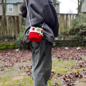 May include: A person wearing a gray hoodie and gray jeans is carrying a small red and white crocheted bag with a green stem and red berries. The bag is attached to a white pearl chain.