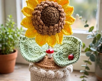 May include: A crocheted sunflower in a beige pot with a textured rim. The flower has bright yellow petals and a brown center. Two green crocheted leaves extend from the base, featuring a small red and white mushroom and a red ladybug.