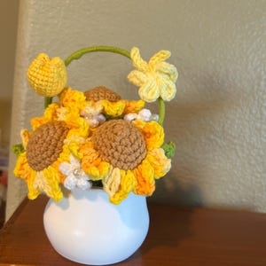 May include: A handcrafted crochet sunflower arrangement in a white vase. The sunflowers have yellow and orange petals with brown centers. A green handle arches over the vase, with a yellow tulip and a yellow flower on the sides. The arrangement sits on a wooden surface.