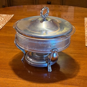 May include: A silver-colored serving dish with a lid and decorative handle. The dish has a detailed, openwork design around the middle and rests on three curved legs. A small bird-shaped detail is on the front.