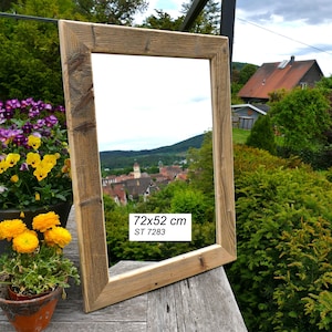 May include: A rectangular mirror with a weathered, light brown wooden frame. The mirror surface reflects a landscape with buildings and trees. A white label on the glass displays the text 72x52 cm and ST 7283 in black, sans-serif font.