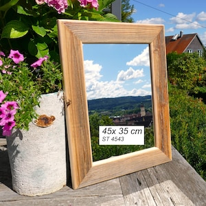 May include: A rectangular mirror with a natural light brown wood frame. The mirror surface reflects a blue sky with clouds and a landscape. A white label on the glass displays the text 45x 35 cm and ST 4543 in black font, indicating the dimensions.
