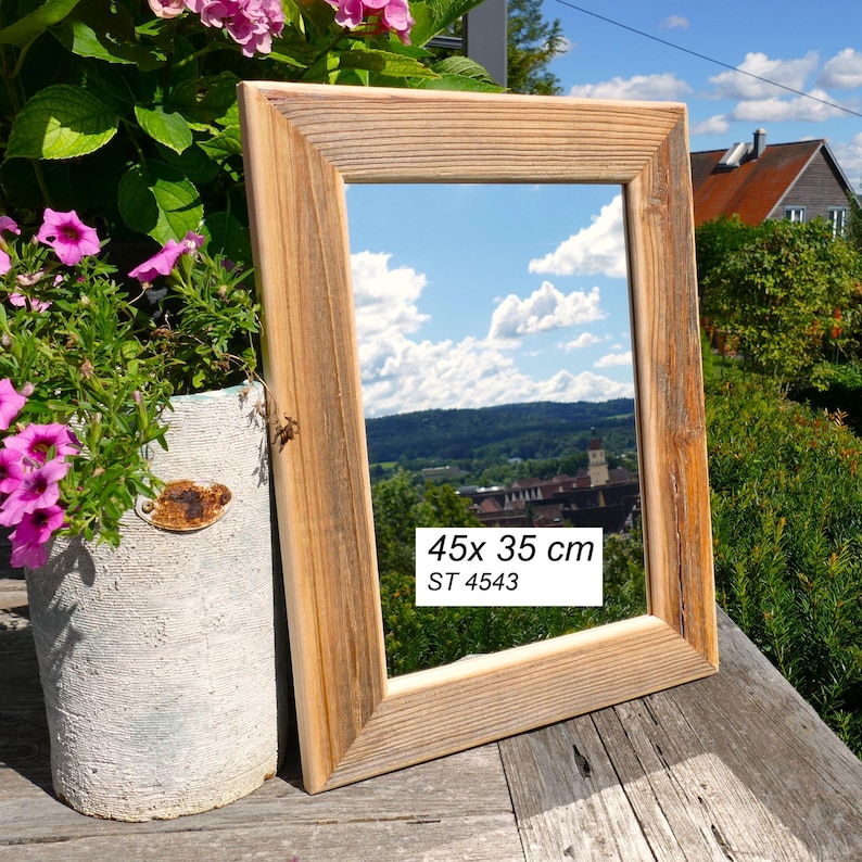 May include: A rectangular mirror with a natural light brown wood frame. The mirror surface reflects a blue sky with clouds and a landscape. A white label on the glass displays the text 45x 35 cm and ST 4543 in black font, indicating the dimensions.
