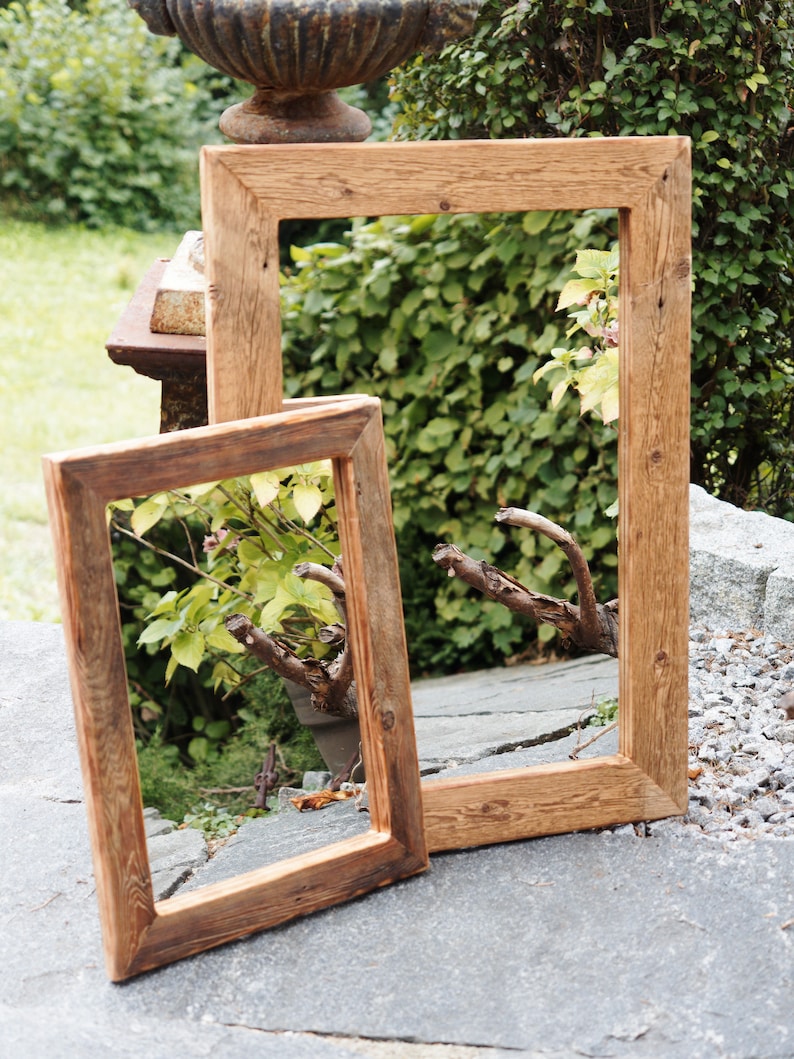 May include: Two rectangular picture frames made of natural brown wood with visible grain and knots. The frames are empty, featuring mitered corners and a simple, flat profile. They are positioned upright, with the smaller frame overlapping the larger one.