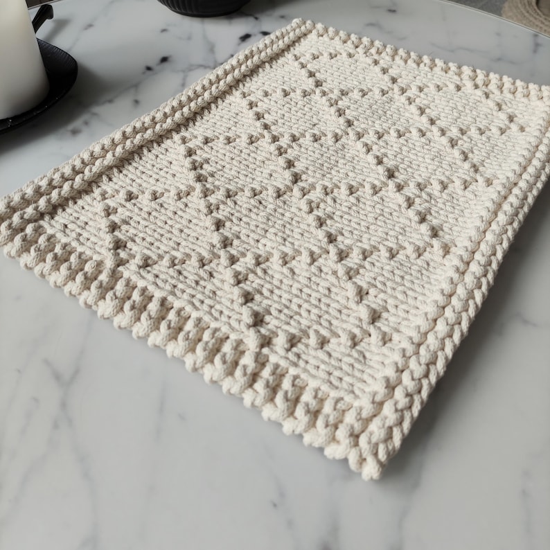 May include: A cream-colored woven cotton placemat with a diamond pattern. The placemat has a textured surface and a fringed edge.