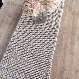 May include: A beige knitted table runner with a rectangular shape, placed on a wooden table. A glass vase with dried pink and cream-colored flowers sits on the runner. The table runner has a small wooden tag.