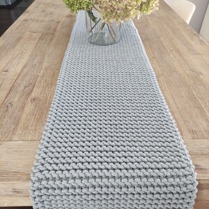 May include: A light grey, knitted table runner is displayed on a wooden table. The runner is rectangular and has a textured, woven appearance. A clear glass vase with green and white flowers sits on the table, behind the runner.