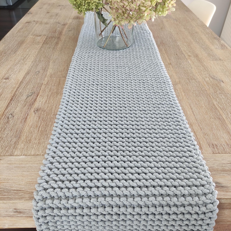 May include: A light grey, knitted table runner is displayed on a wooden table. The runner is rectangular and has a textured, woven appearance. A clear glass vase with green and white flowers sits on the table, behind the runner.