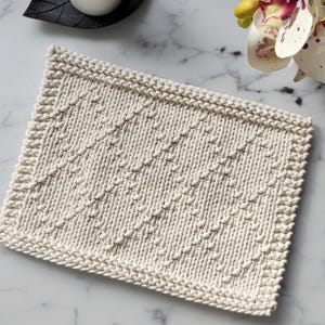 May include: A cream-colored knitted dishcloth with a diamond pattern. The dishcloth is made with a textured yarn and has a simple, elegant design.