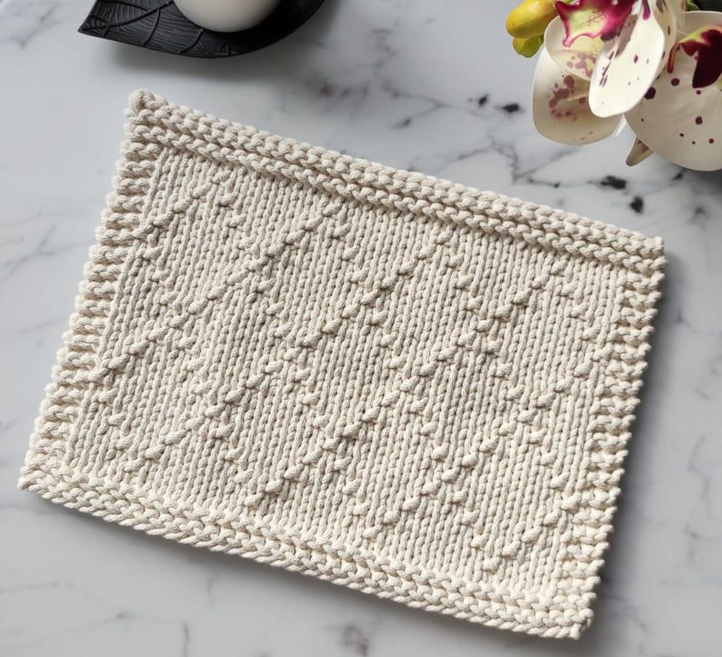 May include: A cream-colored knitted dishcloth with a diamond pattern. The dishcloth is made with a textured yarn and has a simple, elegant design.