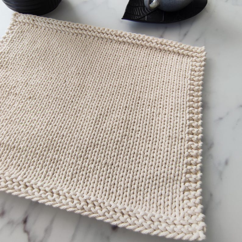 Op de afbeelding: Een rechthoekige, cr&egrave;mekleurige gebreide placemat met een getextureerd oppervlak. De randen hebben een decoratieve rand. De placemat is gemaakt van een dik, zacht garen, geschikt voor keuken- of eetkamerdecoratie.