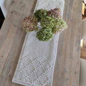 May include: A white knitted table runner with a diamond pattern, featuring a bouquet of pink and green hydrangeas in a clear vase.