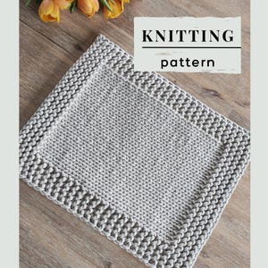 Peut inclure: Un tapis rectangulaire en tricot gris clair avec une bordure texturée et un panneau central lisse. Le texte KNITTING pattern est affiché en lettres noires sur un encart rectangulaire blanc situé en haut de l'image.