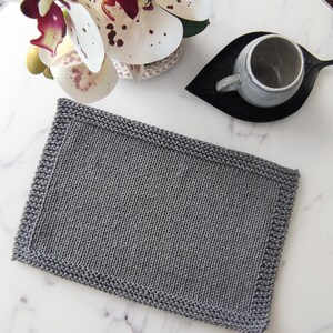 May include: A rectangular, gray knitted placemat with a textured surface and a detailed border. The placemat is shown on a white marble surface, with a gray mug on a black leaf-shaped coaster and a white and purple orchid in the background.