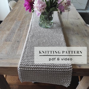 May include: A light beige knitted table runner with a textured stitch pattern. A clear glass vase containing pink and white peonies sits on the runner. The text KNITTING PATTERN pdf & video is overlaid on the image in black lettering.