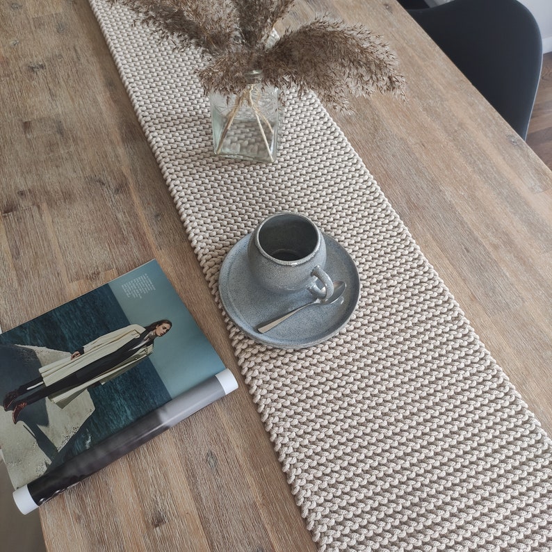 May include: A beige knitted table runner with a textured pattern, on a wooden table. A grey ceramic teacup and saucer with a spoon are on the runner. A magazine and a glass vase with dried plants complete the scene.