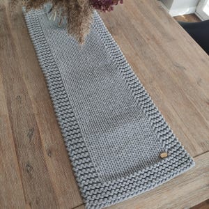 Könnte beinhalten: Ein rechteckiger, hellgrauer Tischläufer mit grober Strickstruktur und einem markanten Randmuster. Der Tischläufer ist in der unteren rechten Ecke mit einem kleinen, ovalen Holzschild versehen, das einen Kontrast zum grauen Garn bildet.
