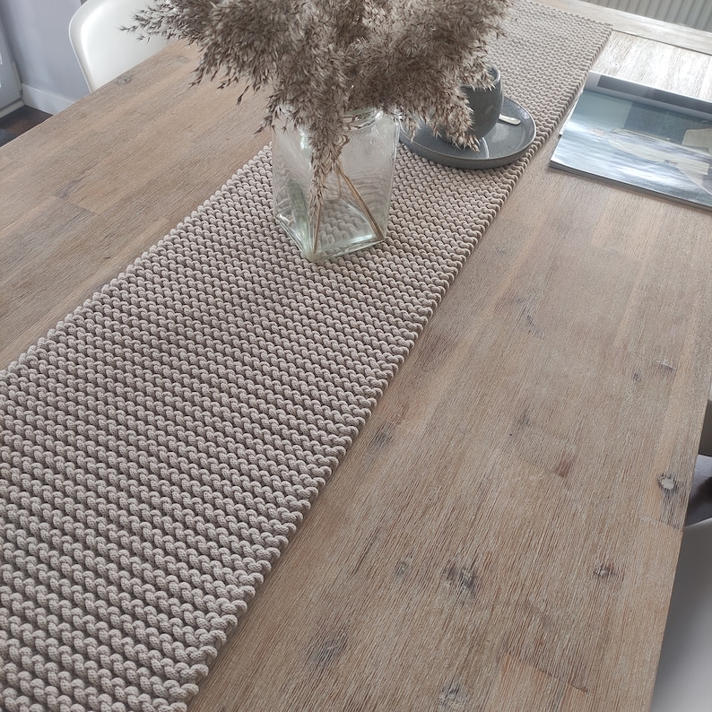 May include: A beige knitted table runner is placed on a light brown wooden table. A clear glass vase with dried plants sits on the runner, along with a grey saucer and a cup. A magazine is on the right side of the table.