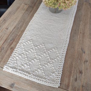 May include: A white knitted table runner with a textured pattern. The runner is laid out on a wooden table.